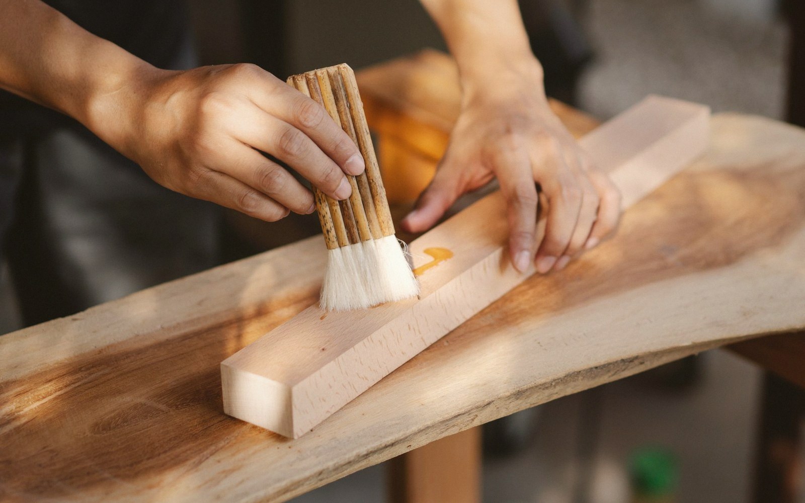 Hands apply oil with a brush to a wooden block on a live-edge table