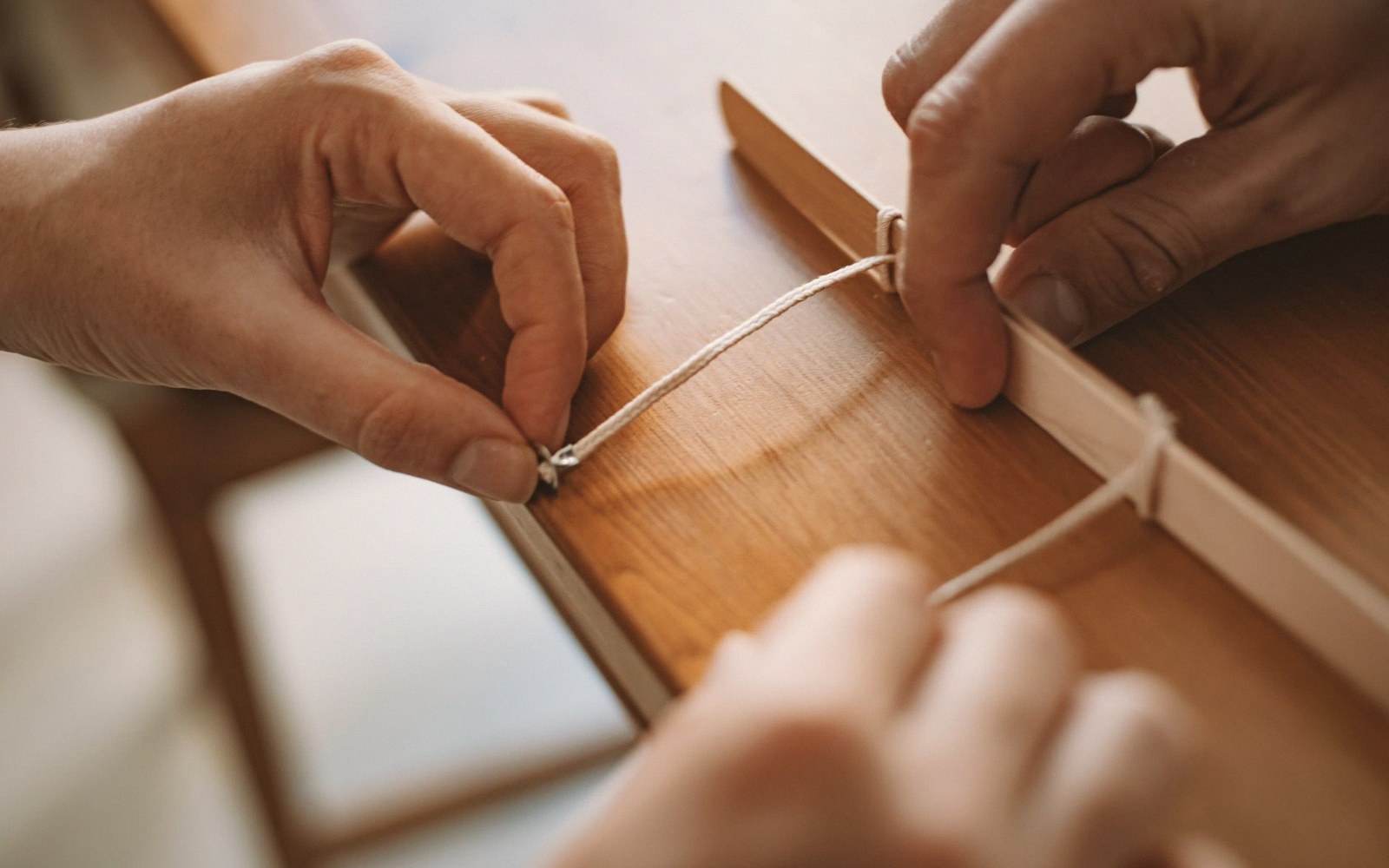 Two hands tying a cord knot along the edge of a wooden panel